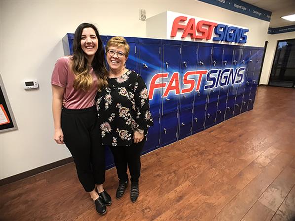 Emily Shuler and Susan Shea smile together next to FASTSIGNS lockers